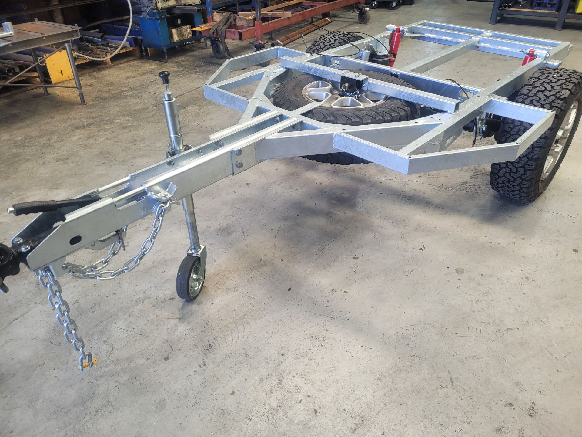 A galvanized steel trailer frame with a mounted spare tire and hitch, parked on a concrete workshop floor.