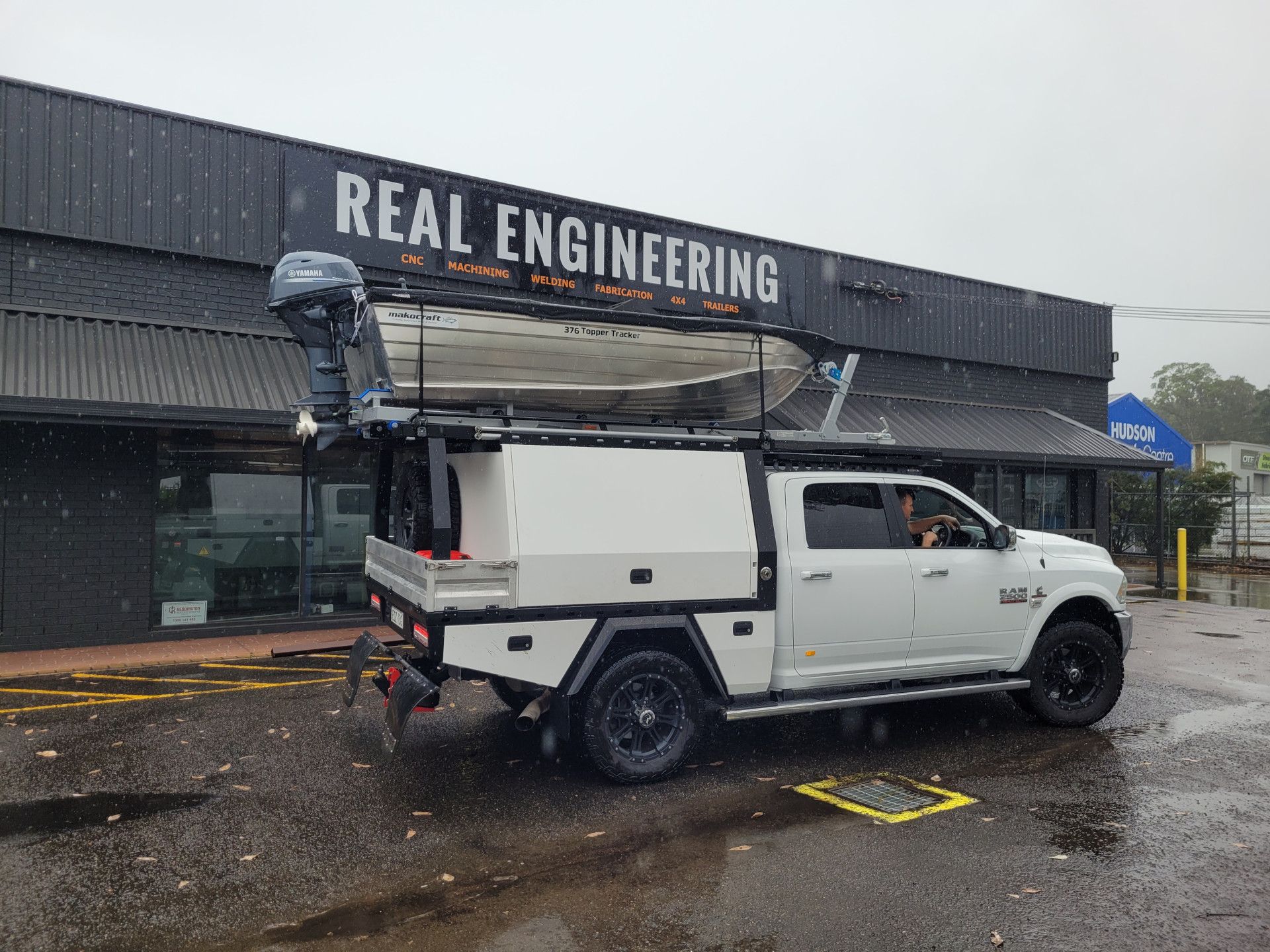 A white pickup truck with a utility canopy and a small boat with an outboard motor mounted on its roof rack.