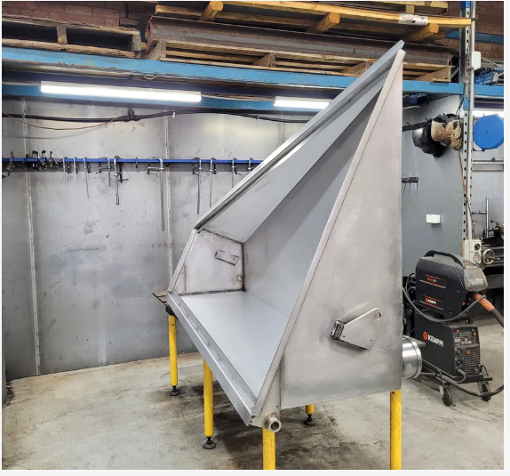 Large gray metal hopper or chute in an industrial workshop, mounted on yellow legs under overhead piping. — Real Engineering in West Gosford, NSW