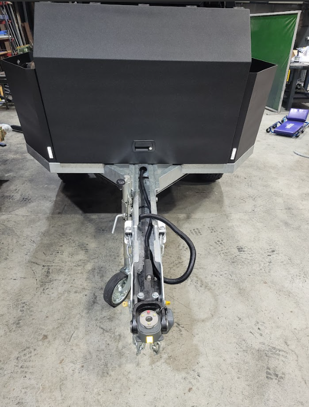 A matte black trailer tongue and hitch assembly extending from a workshop floor, viewed from a high angle.