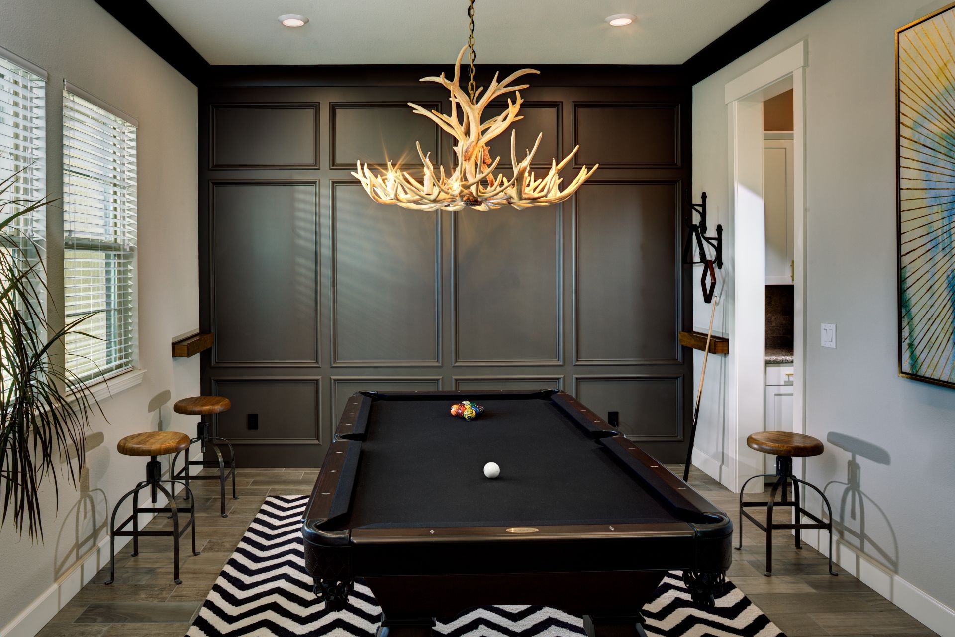 Pool table in a room with dark paneled wall, antler chandelier, and patterned rug.