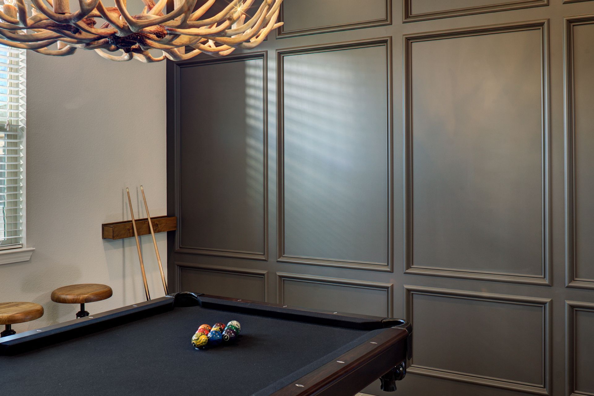 Pool table in room with antler chandelier, wainscoting, stools, pool cues, and balls.