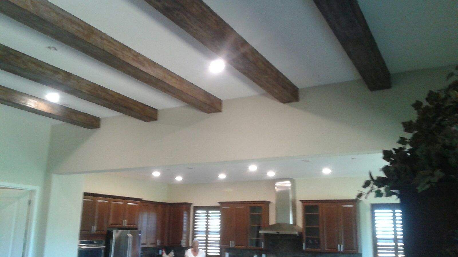 Wooden beams on a vaulted ceiling in a kitchen with recessed lighting, dark cabinets, and a stainless steel range hood.