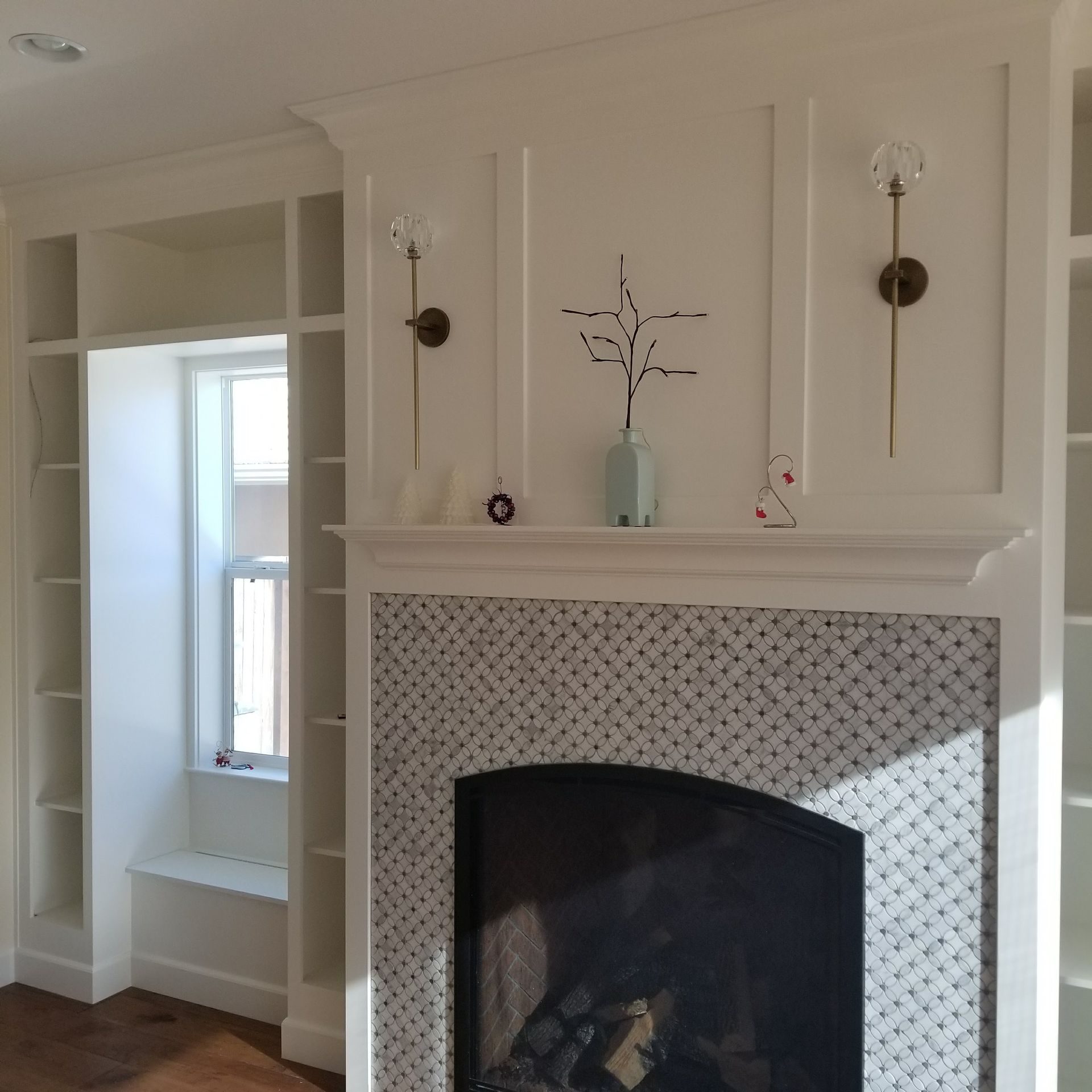 Built-in white bookshelves flank a fireplace with patterned tile surround. Two sconces and a decorative vase sit above the hearth.