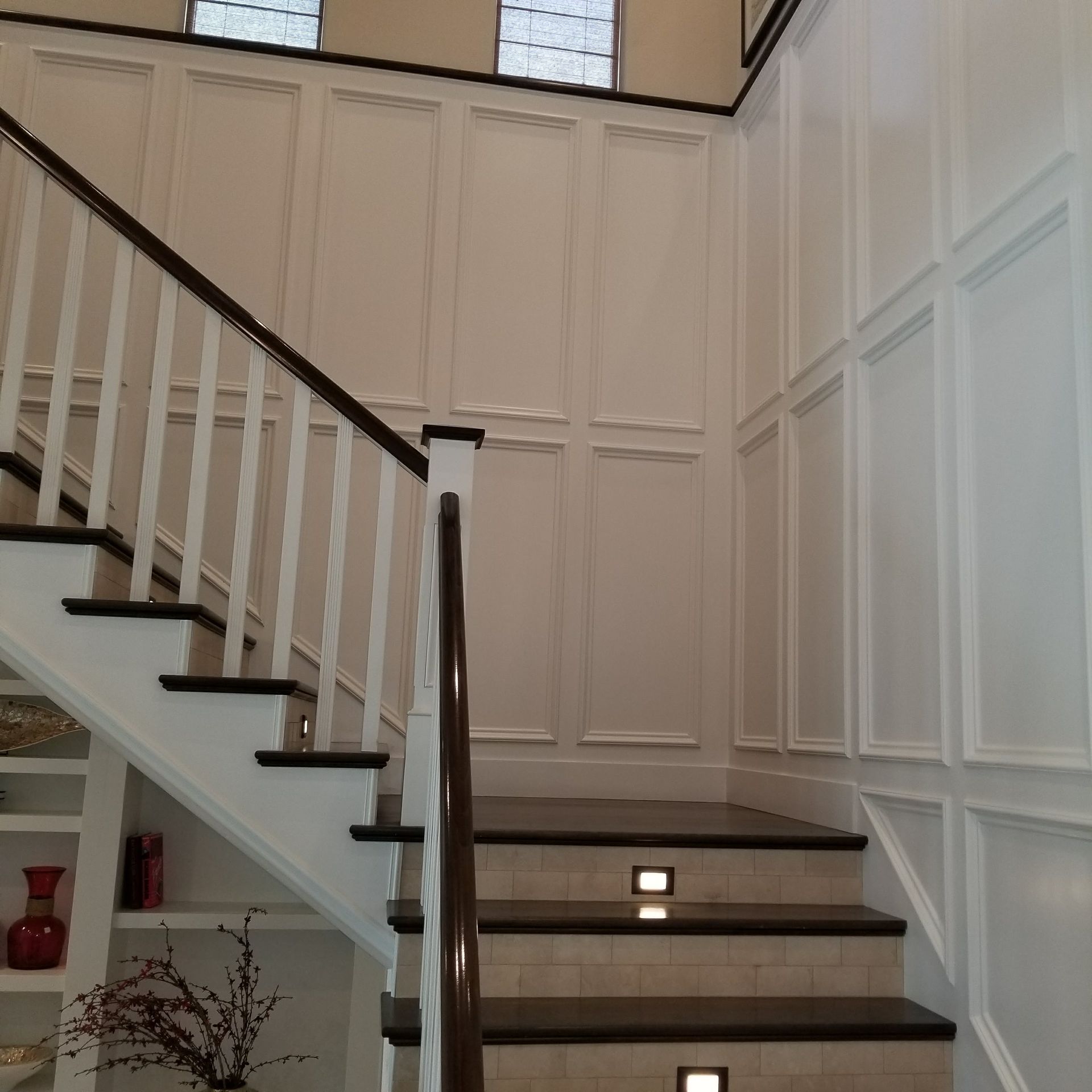 Staircase with white paneled walls and dark wooden handrail, leading upwards, lit by recessed lights.
