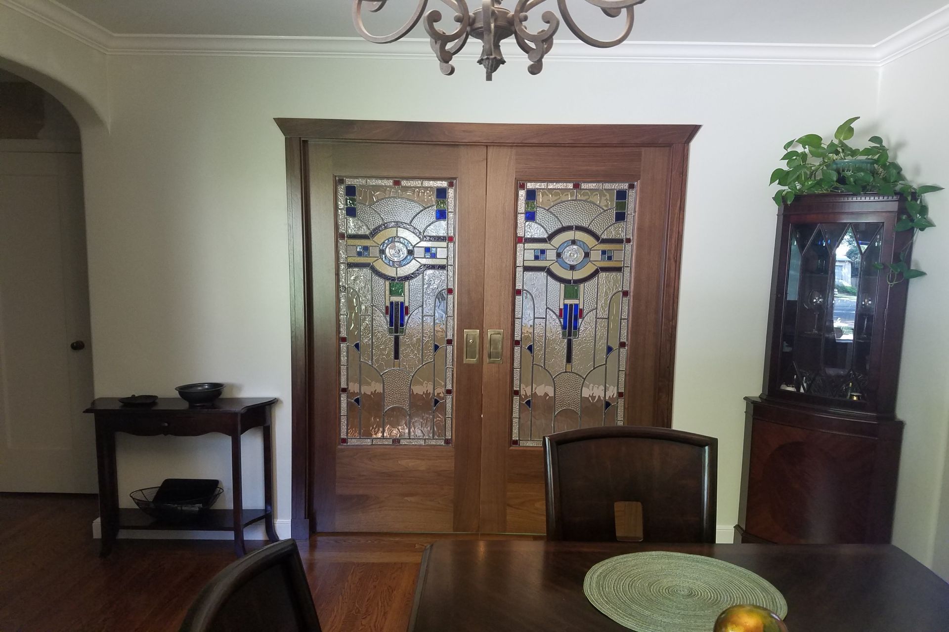 Wooden double doors with stained glass panels, a dining table, and a cabinet in a room.
