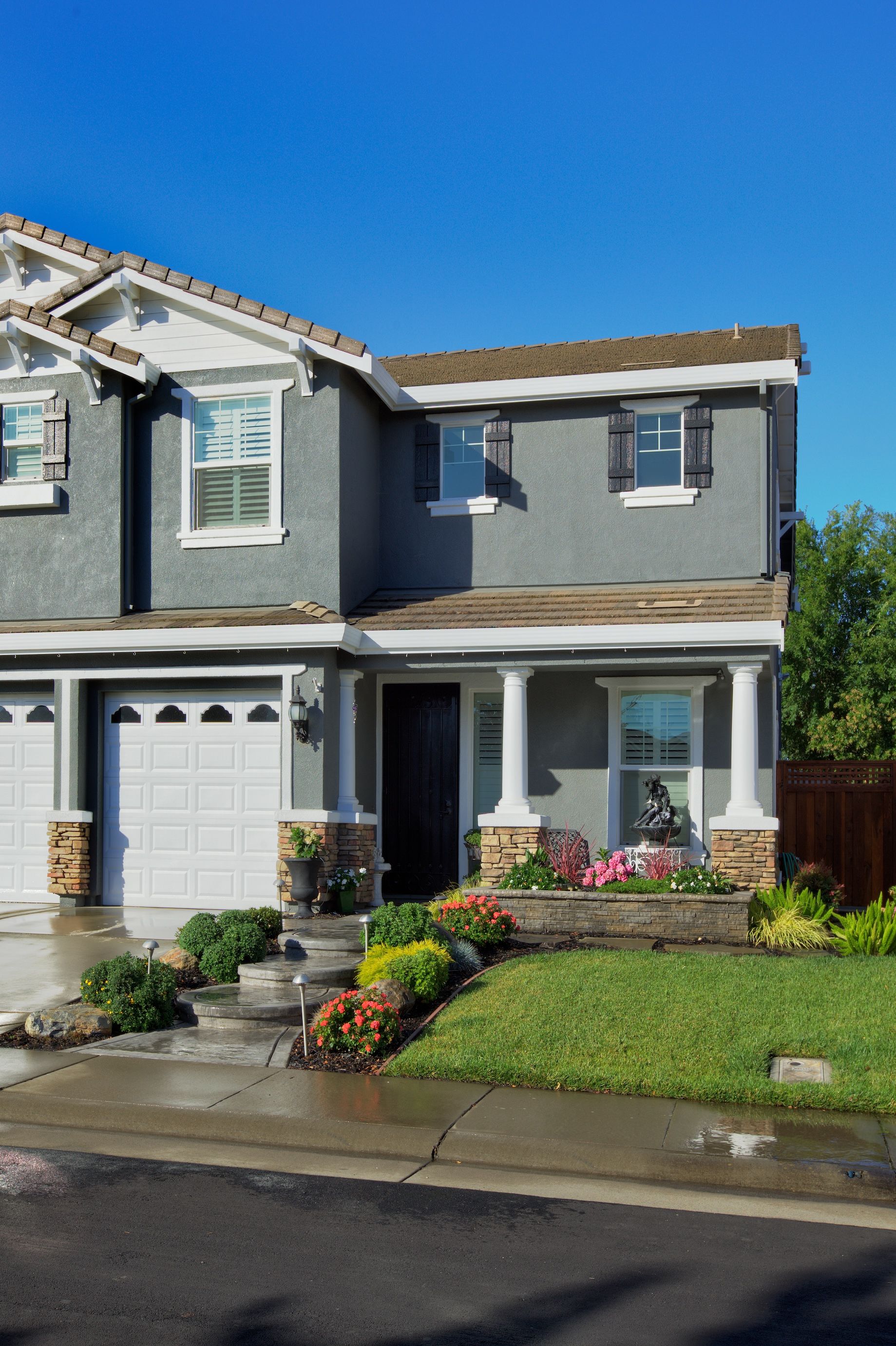Two-story gray house with white trim, flower beds, and a blue sky.