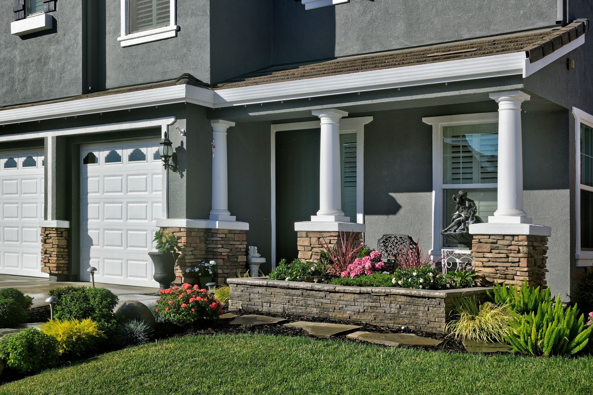 Gray house with white trim, columns, and garage doors. Landscaped garden with green grass and colorful flowers.
