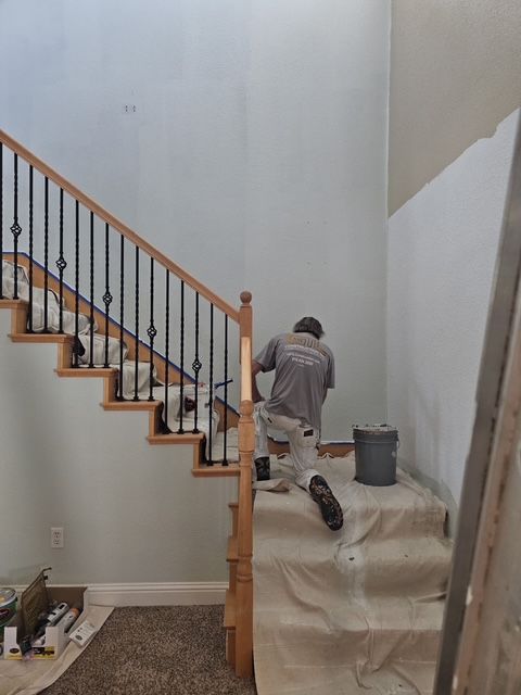 A person painting a staircase, with a wooden banister and black spindles. The walls are pale blue.