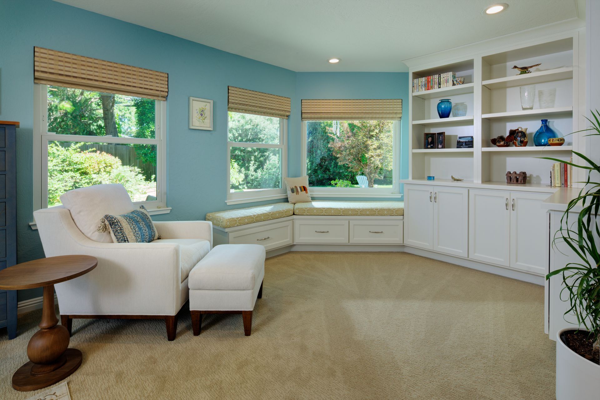 A sunny living room with blue walls, white armchair, and built-in bookshelves, a window seat, and tan carpet.