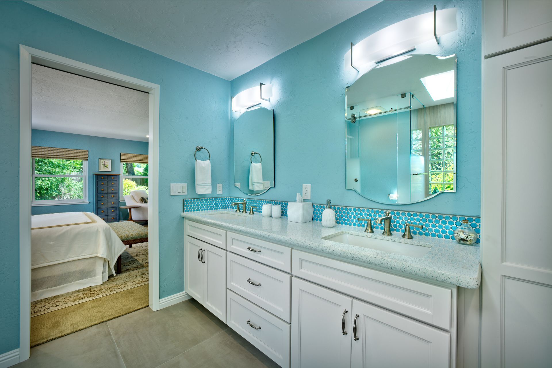 Blue bathroom with double sinks, white cabinets, and a view of a bedroom.