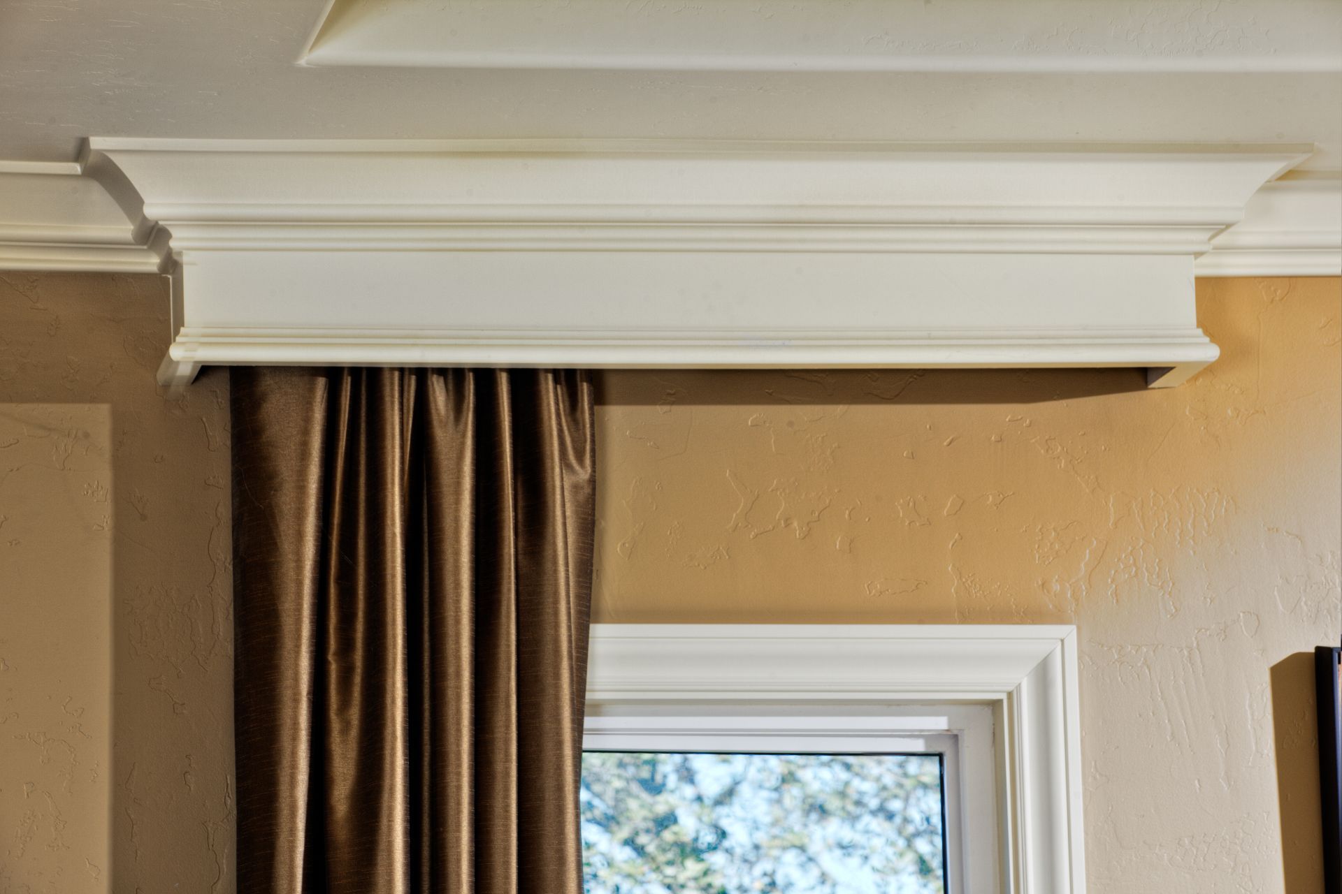 Cream-colored decorative crown molding above a window with a brown curtain and light tan wall.