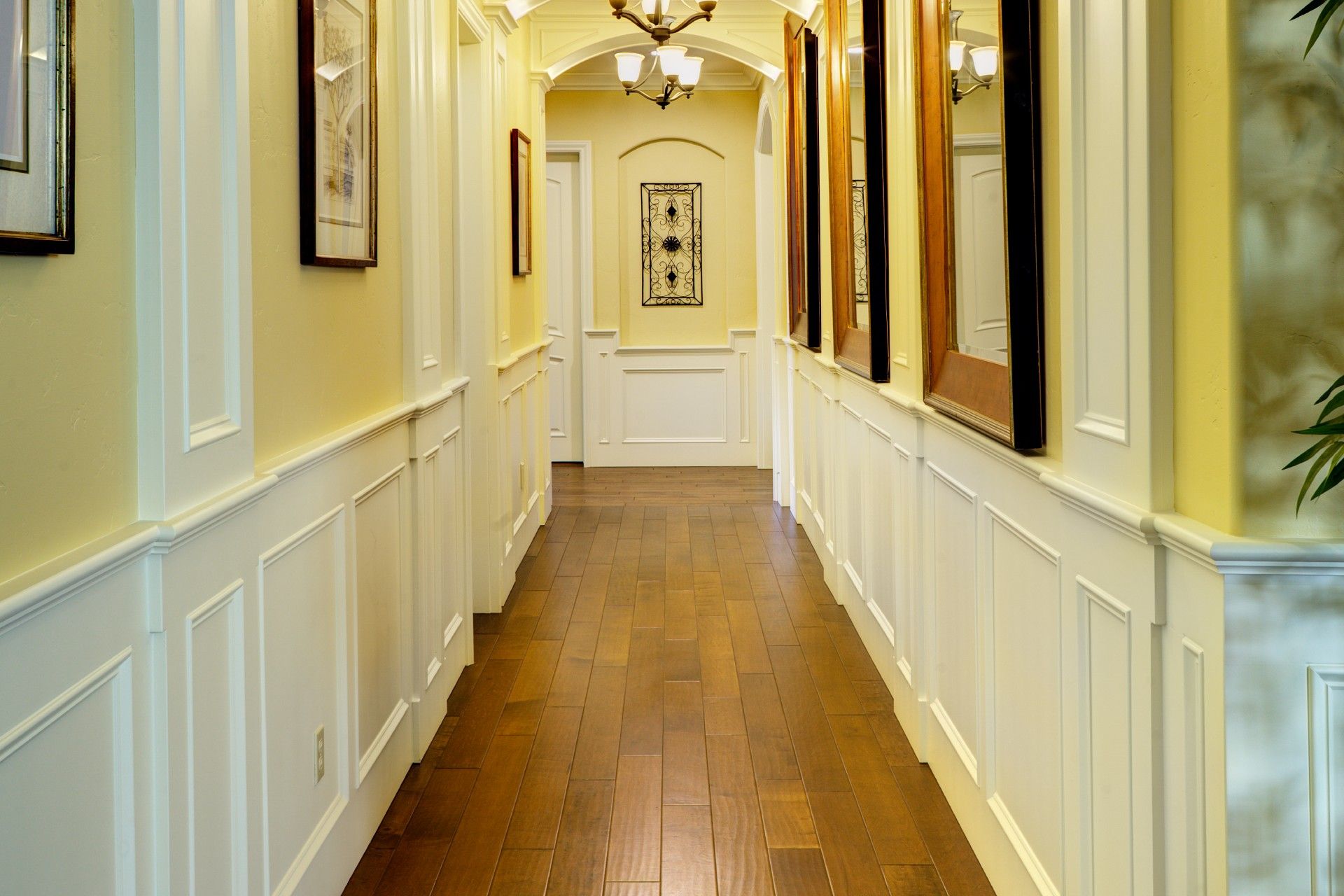 Long hallway with wood floor, white wainscoting, and yellow walls, leading to an arched doorway.