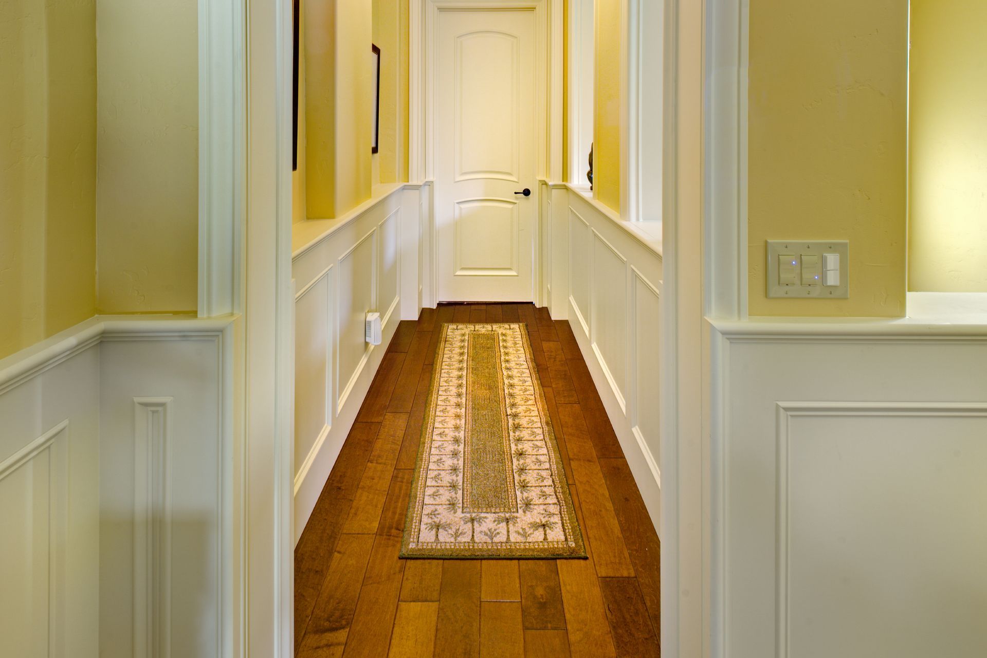 Hallway with wood floor, cream wainscoting, yellow walls, and a patterned rug. Door at the end.