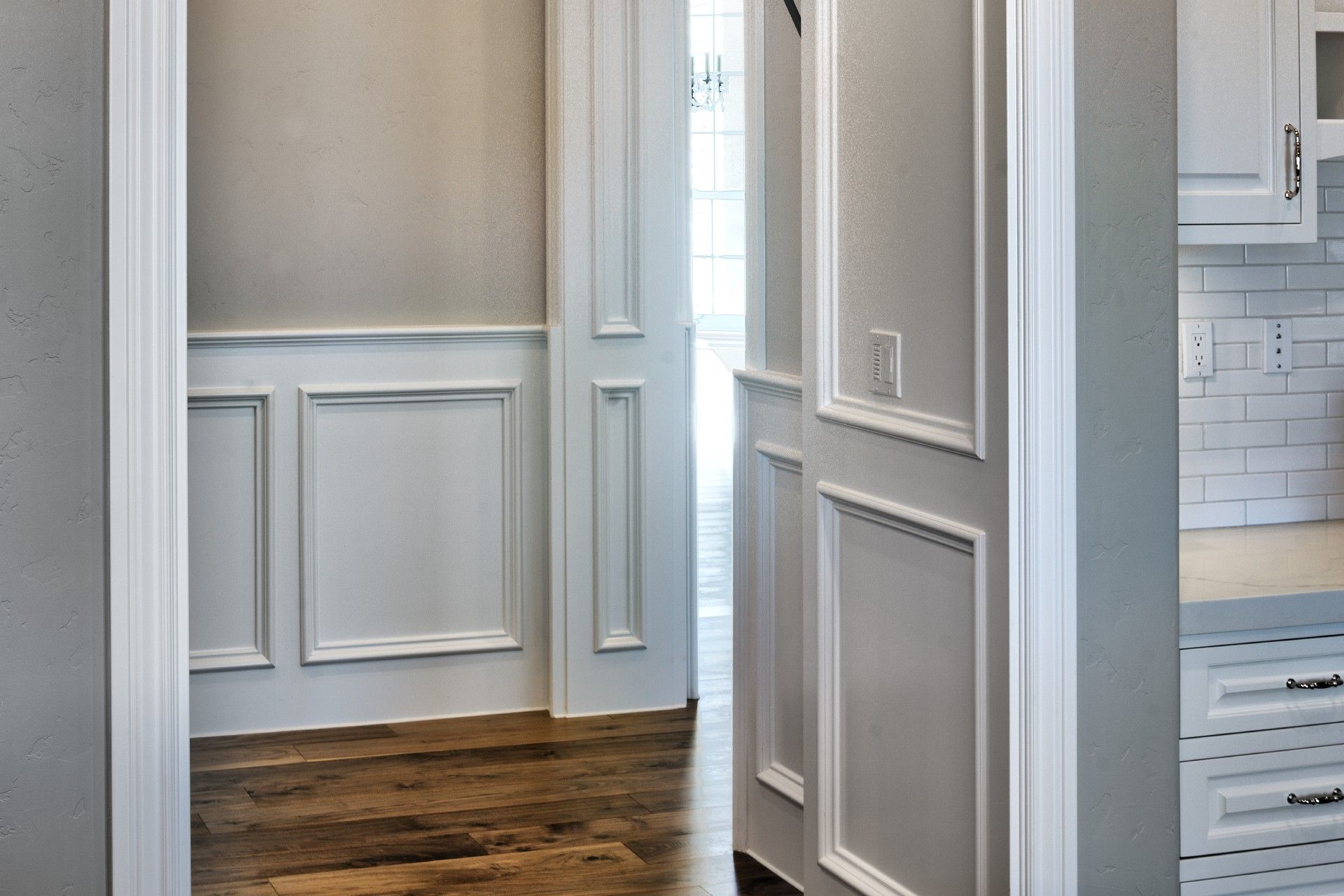 Hallway with white paneled walls, dark wood floors, and an open doorway to a kitchen.