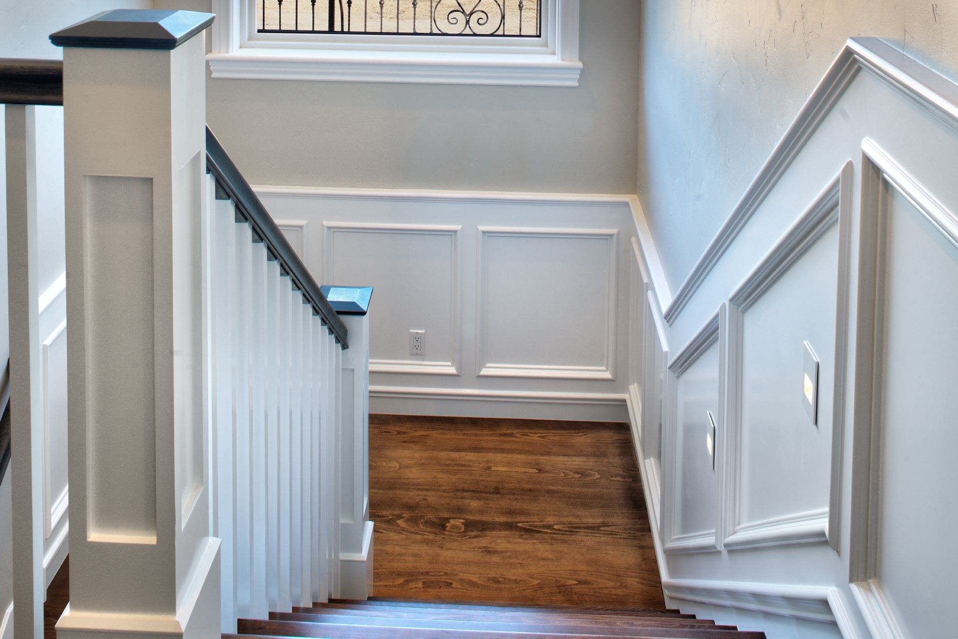 Stairwell with white paneled walls, dark railing, wooden stairs.