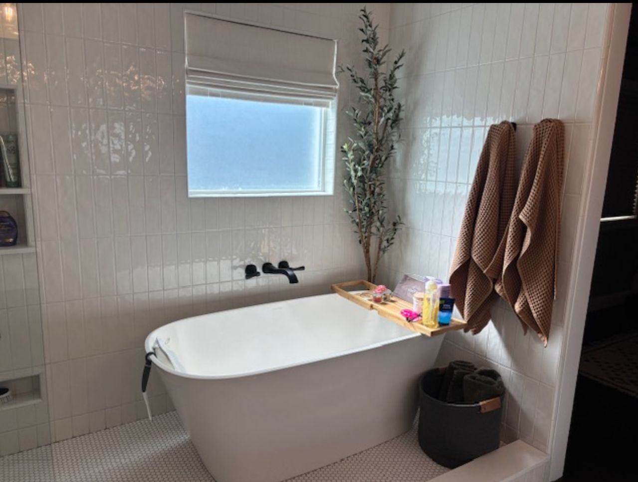 White bathroom with a freestanding tub, white tile, window, towels, and a plant.