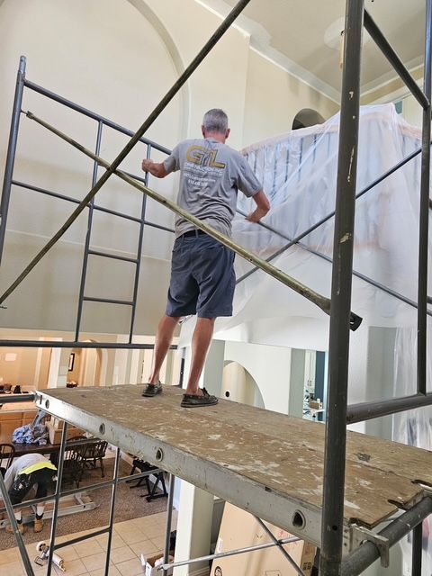 Man on scaffolding near a white-covered object; inside a building with arches, high ceiling, and pews.