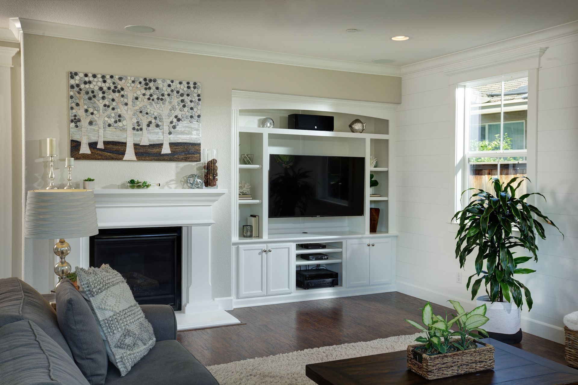 Living room with white built-in entertainment center, fireplace, and plant. Gray sofa and artwork.