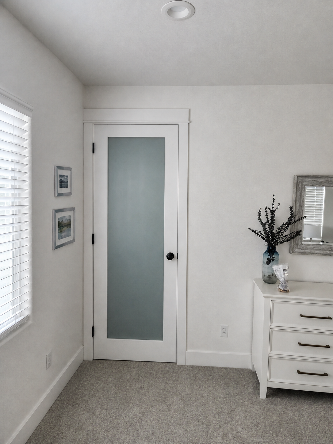 Bright bedroom corner with white door, dresser, window blinds, and beige carpet.
