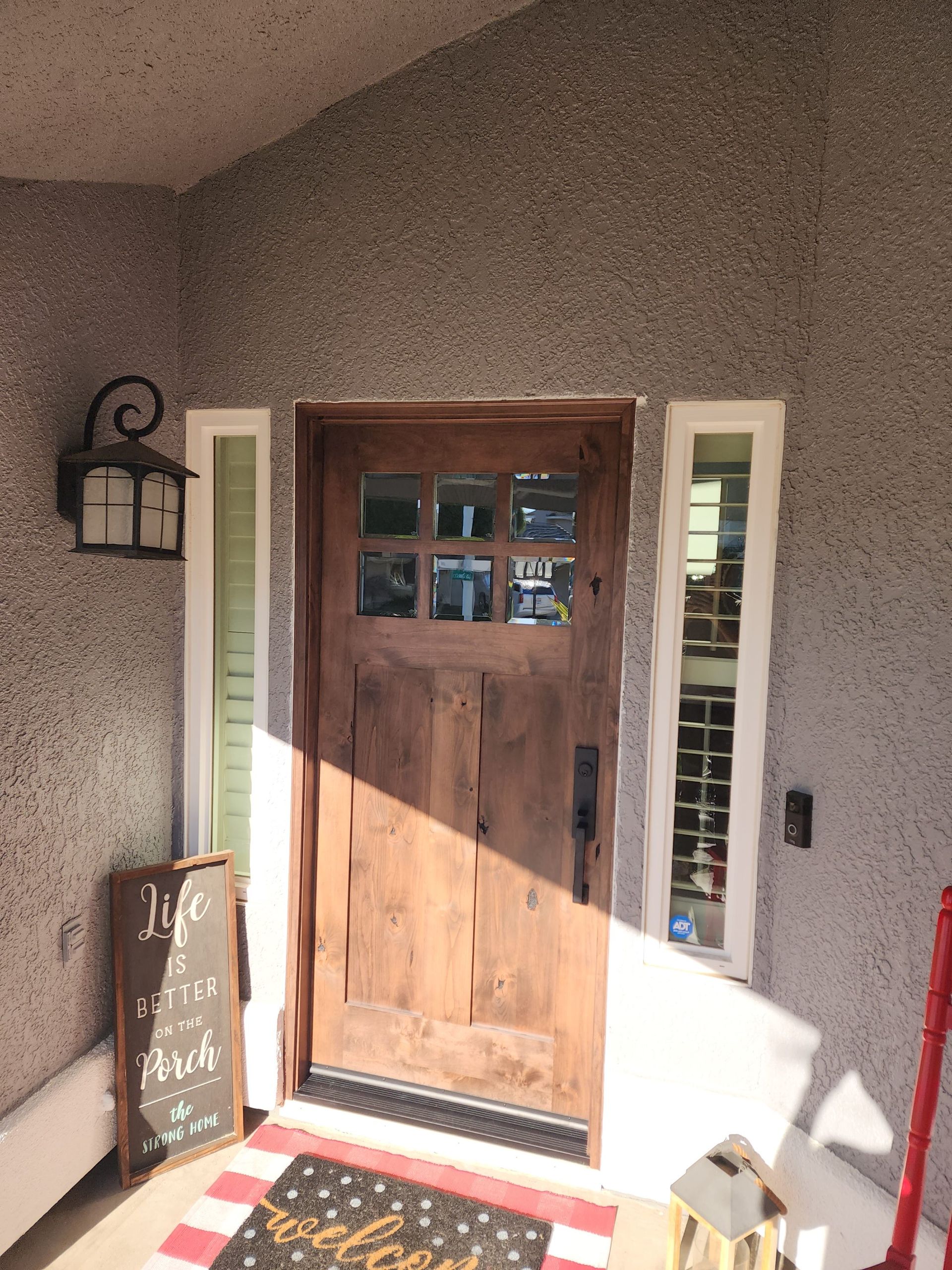 A wooden front door with a windowed top panel, flanked by narrow vertical windows, beside a wall lantern and welcome mat.