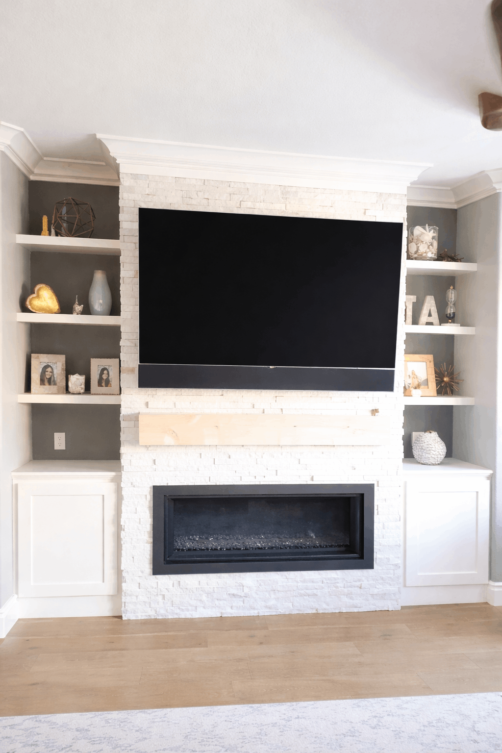 Built-in entertainment center with fireplace. White shelves flank a large TV above a black fireplace, all on a light wood floor.