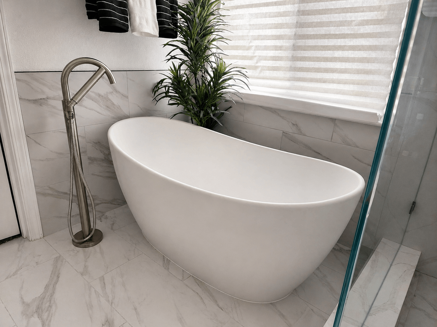 Modern white freestanding bathtub in a bright marble-tiled bathroom next to a floor faucet