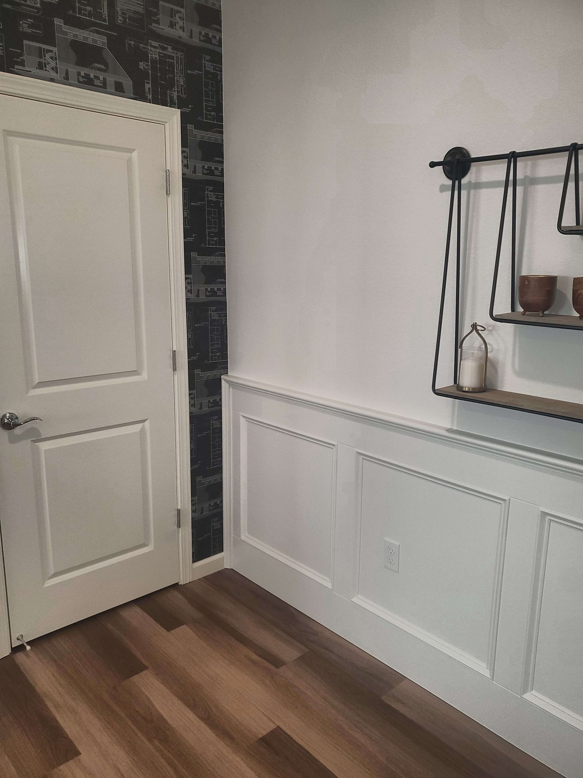 Interior with white wainscoting, wood floor, door, and a black and white accent wall. Shelf with decor.