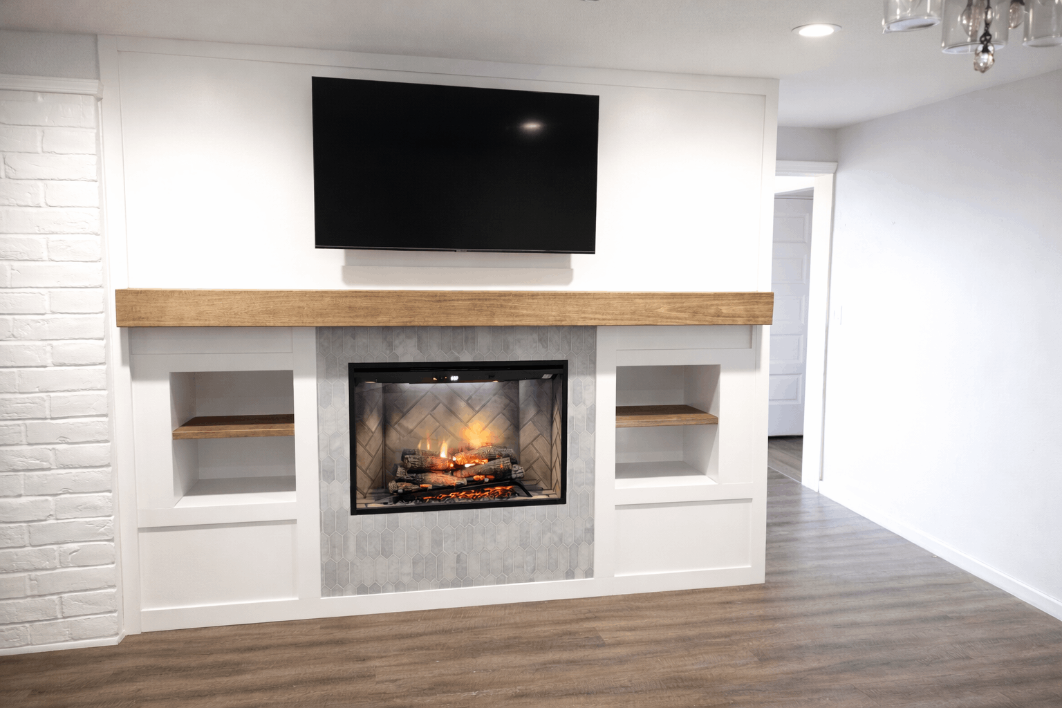 Living room with fireplace, TV above. White walls, wooden mantle, shelves, and brick accents.