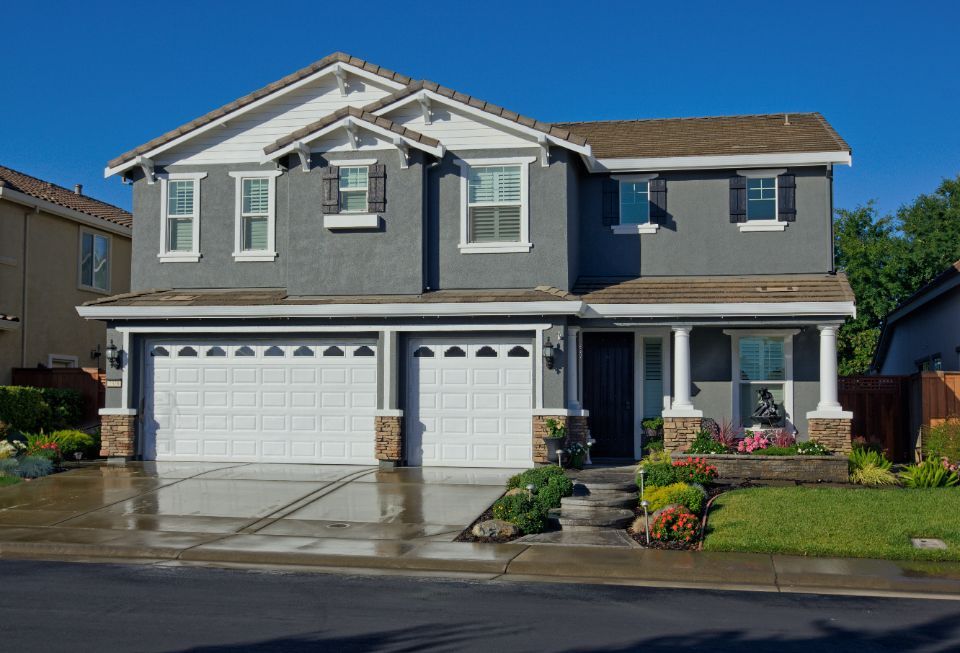 Two-story gray house with white trim, two-car garage, and well-kept lawn on a sunny day.