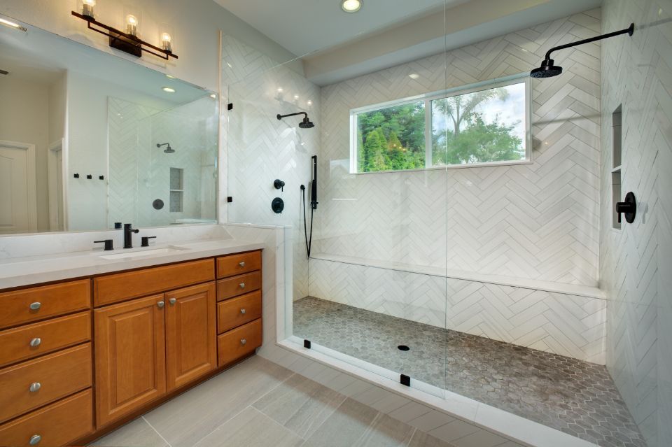 Bathroom with light wood cabinets, white tile herringbone shower, and large window.