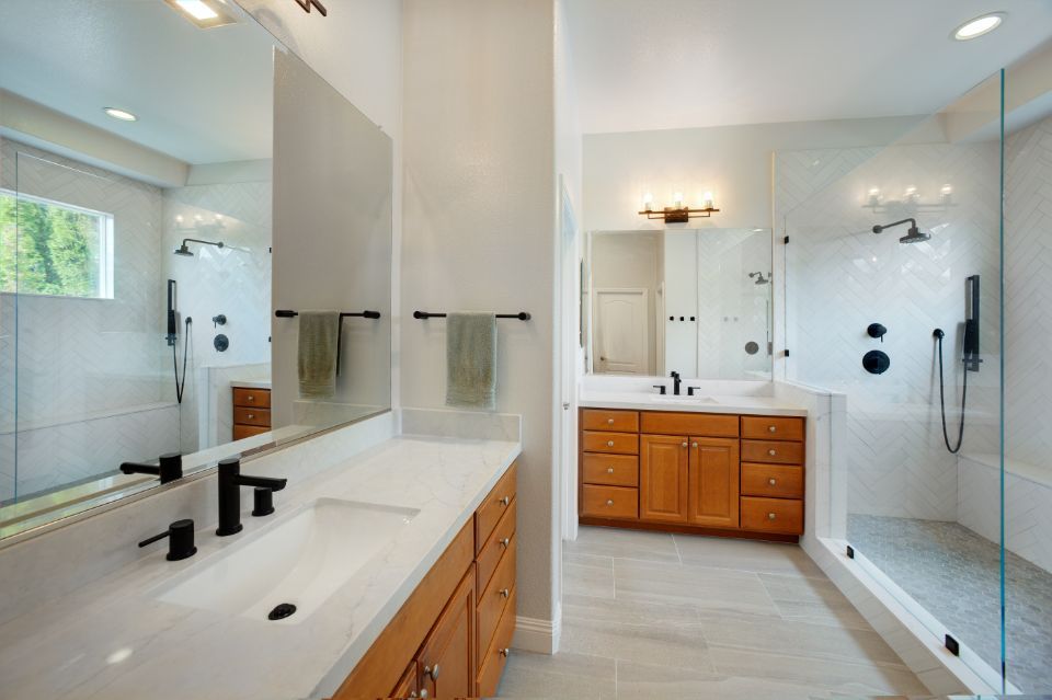 Modern bathroom with double vanity, wooden cabinets, glass shower, white tile, and dark fixtures.