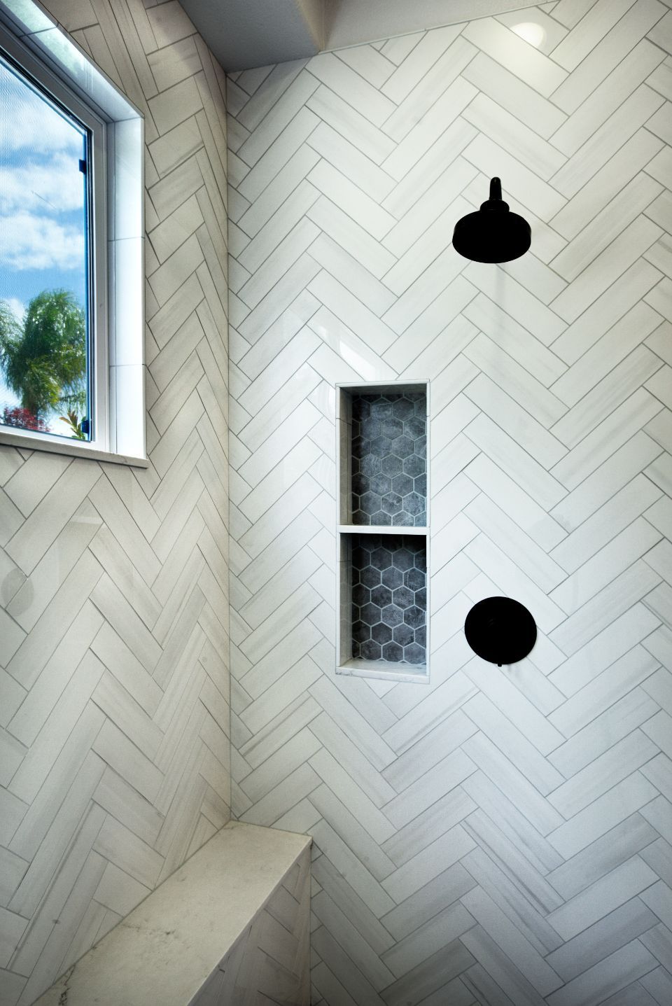 A modern white tiled shower with black fixtures, a bench, and a window with a view of trees and blue sky.