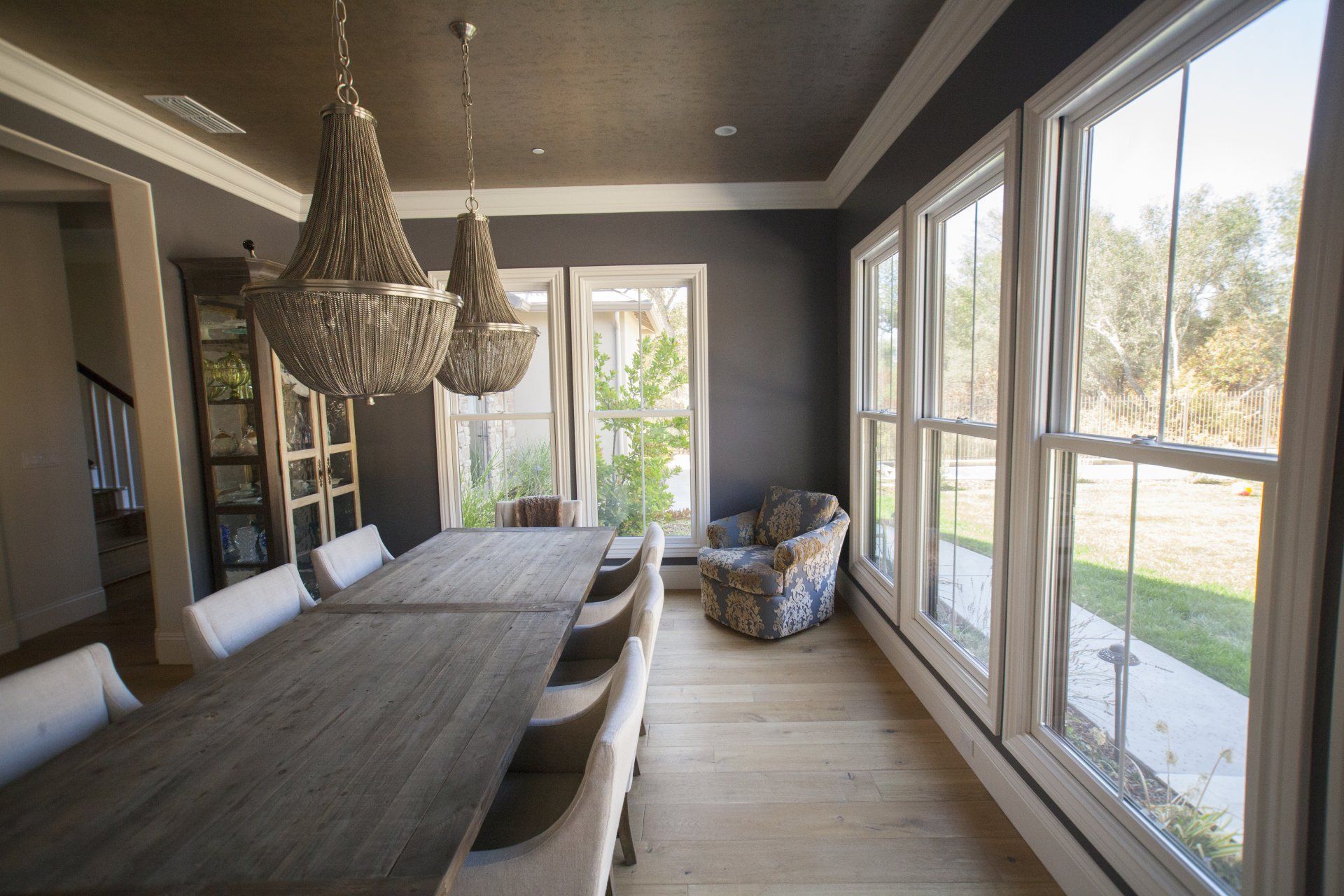 Dining room with long table, chandeliers, windows, gray walls, and a patterned armchair.