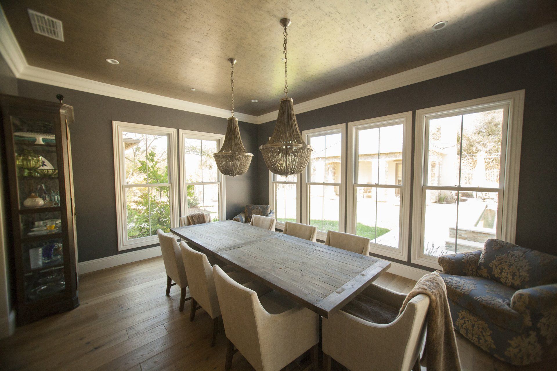 Dining room with long wooden table, chairs, chandeliers, large windows, and a cozy armchair.