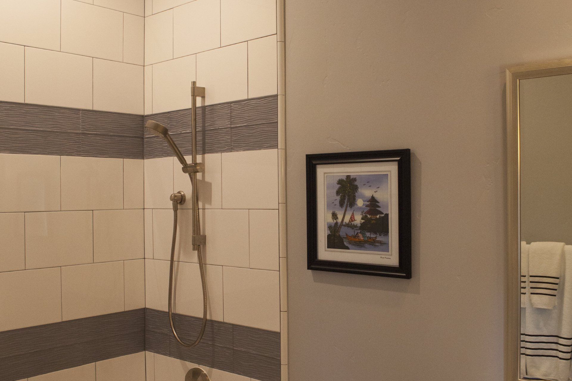 Bathroom with a tiled shower, shower head, and framed artwork on the wall. A mirror with towels is to the right.