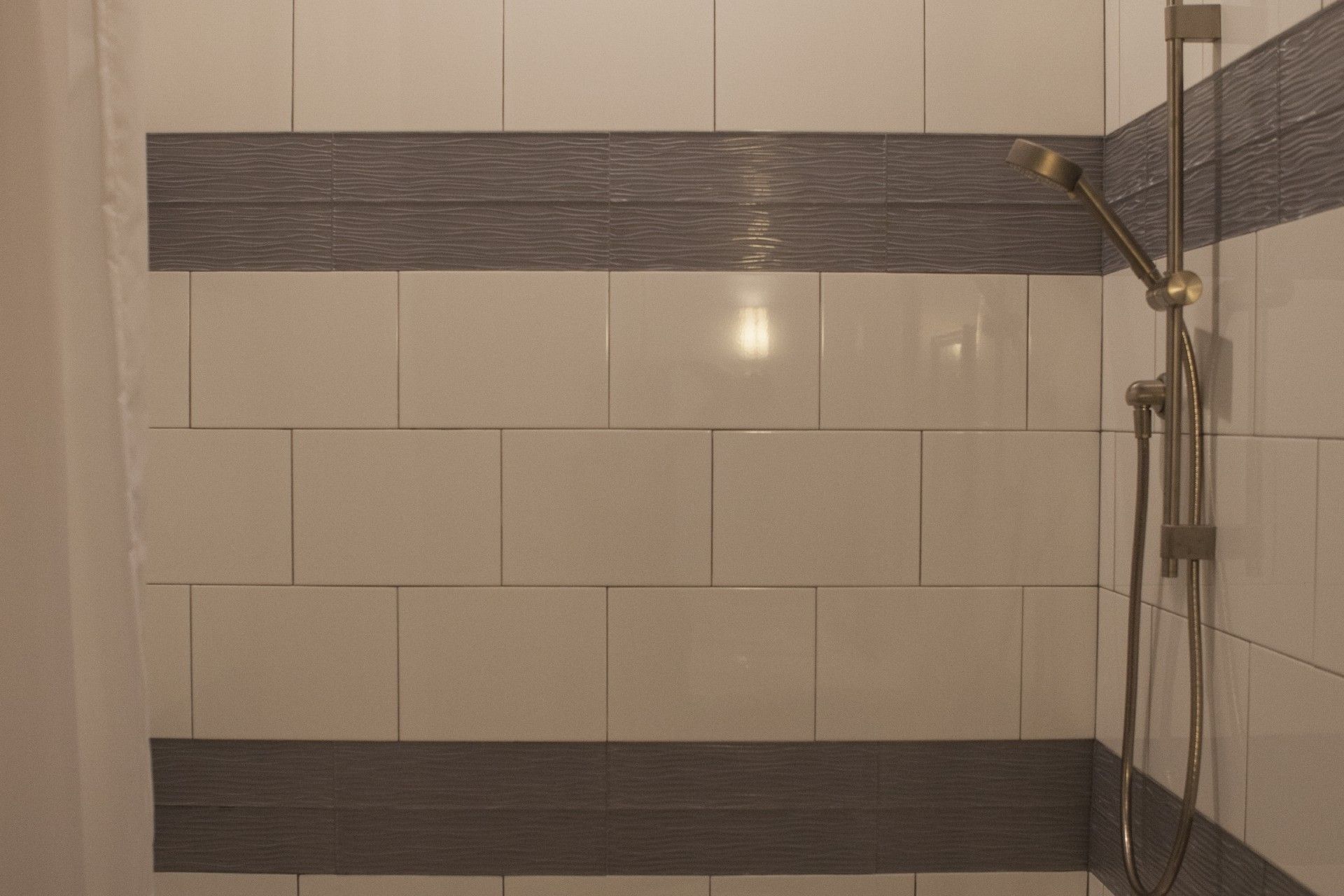 Shower stall with white and gray tiles, a golden shower head, and plumbing.