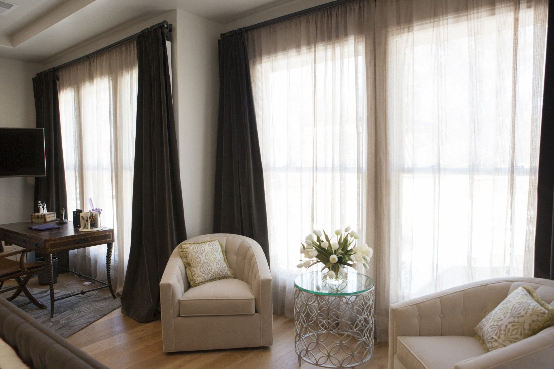 Cozy room with sheer white curtains, dark drapes, armchairs, a small desk, and a glass table with flowers.