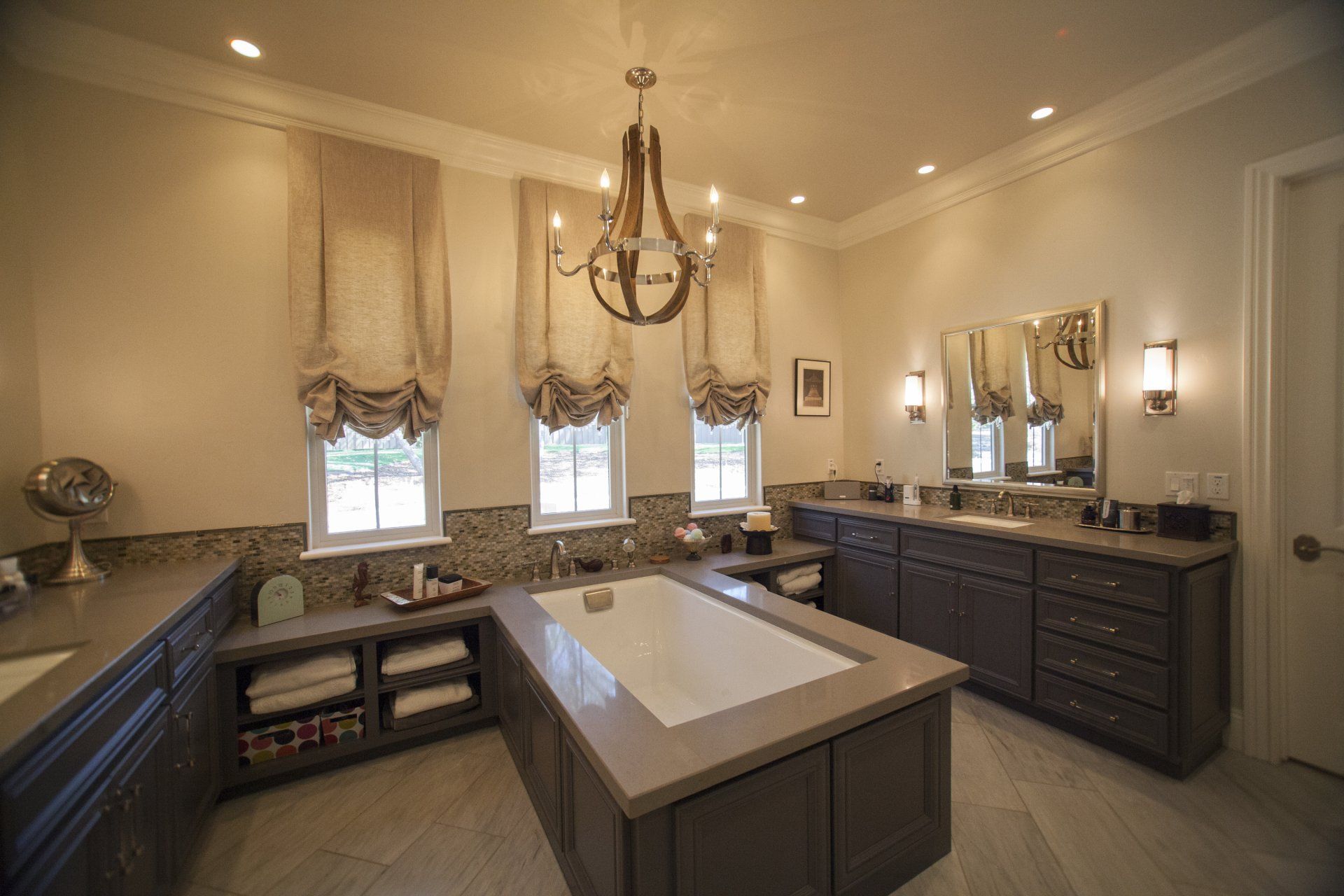 Spacious luxury bathroom with center tub, double sinks, chandelier, windows with curtains, and dark brown cabinets.