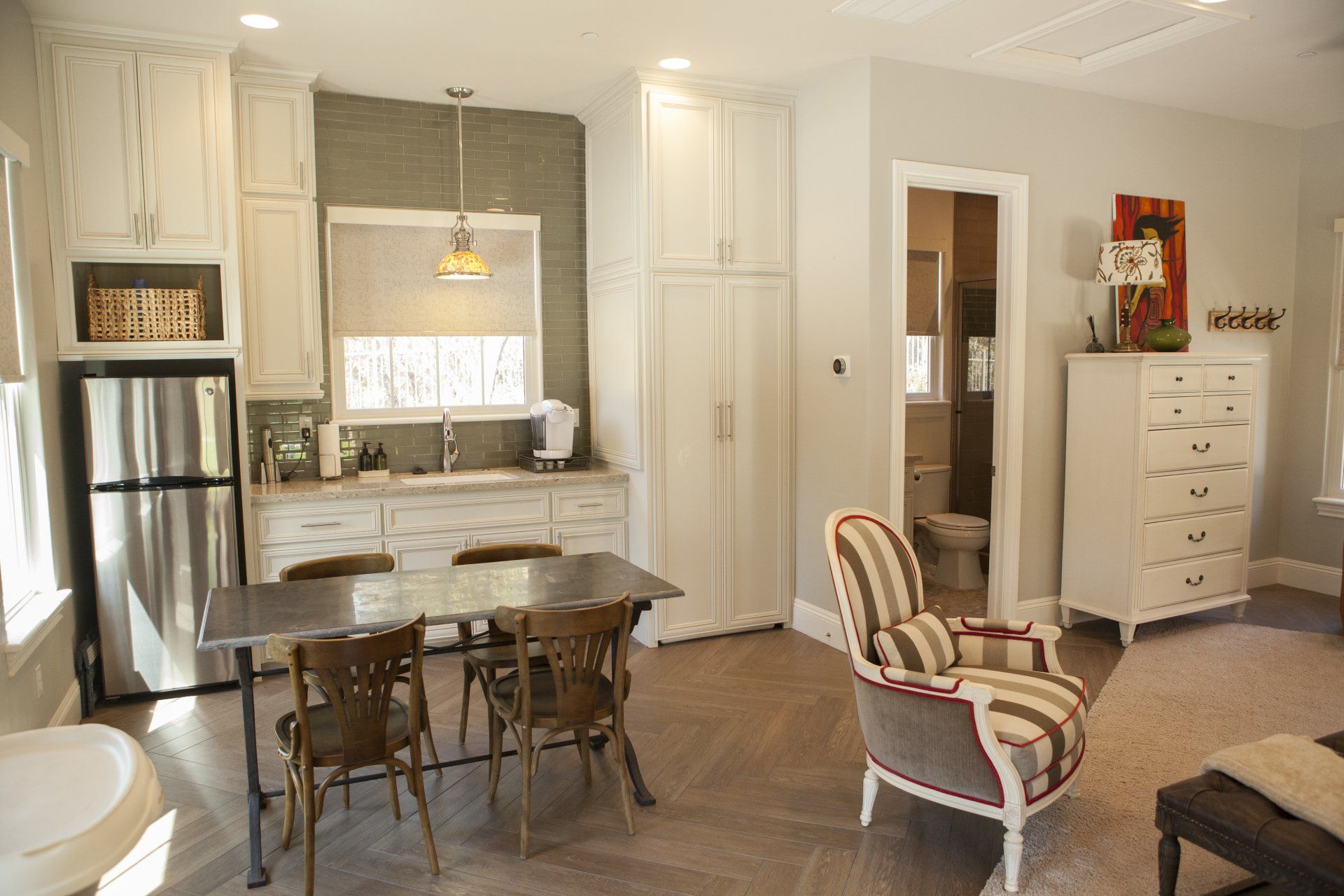 Cozy kitchen and dining area: stainless steel fridge, white cabinets, table with chairs, accent chair near open bathroom.