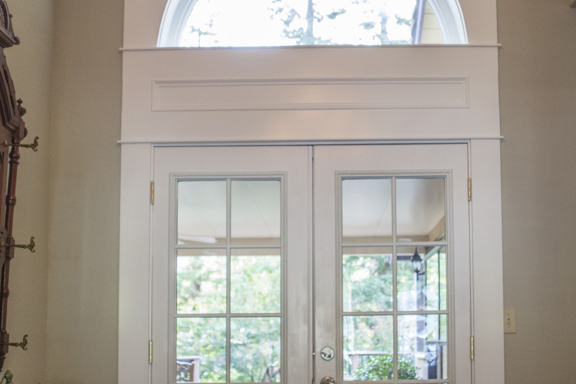 White double doors with glass panes, topped by an arched window and molding, in a beige-walled entryway.