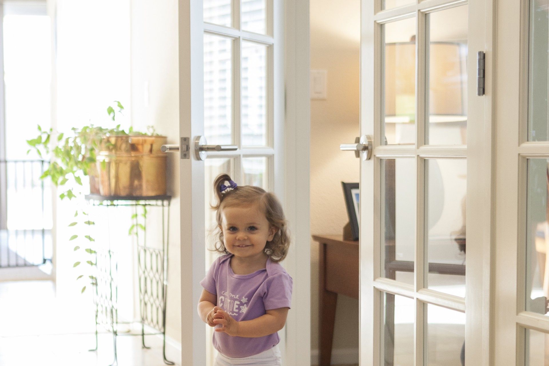 Smiling toddler stands between white French doors, bathed in sunlight.