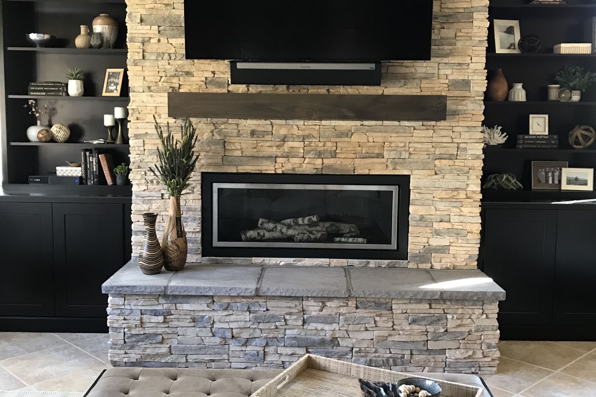 Fireplace with stone surround, built-in black bookshelves, dark mantle, flat screen TV, and decorative vases.