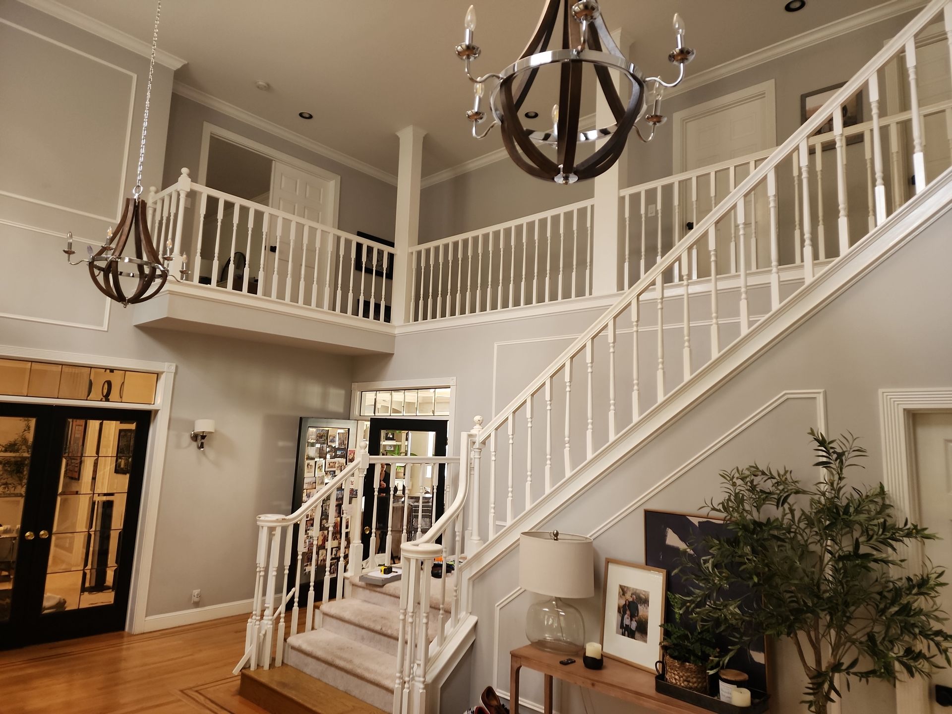 A grand foyer with white staircase, two-story railing, and gray walls. Two chandeliers hang from the ceiling.