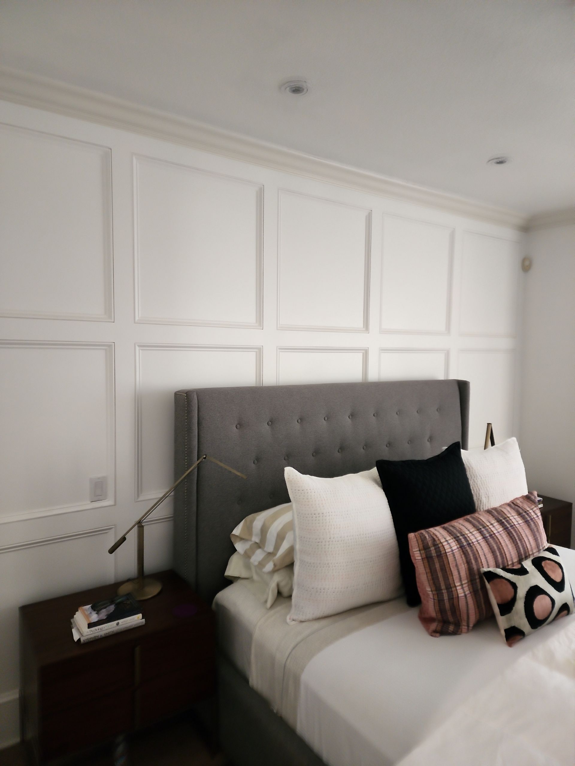 Bedroom with gray upholstered headboard, white paneled walls, dark wood nightstand, and decorative pillows.