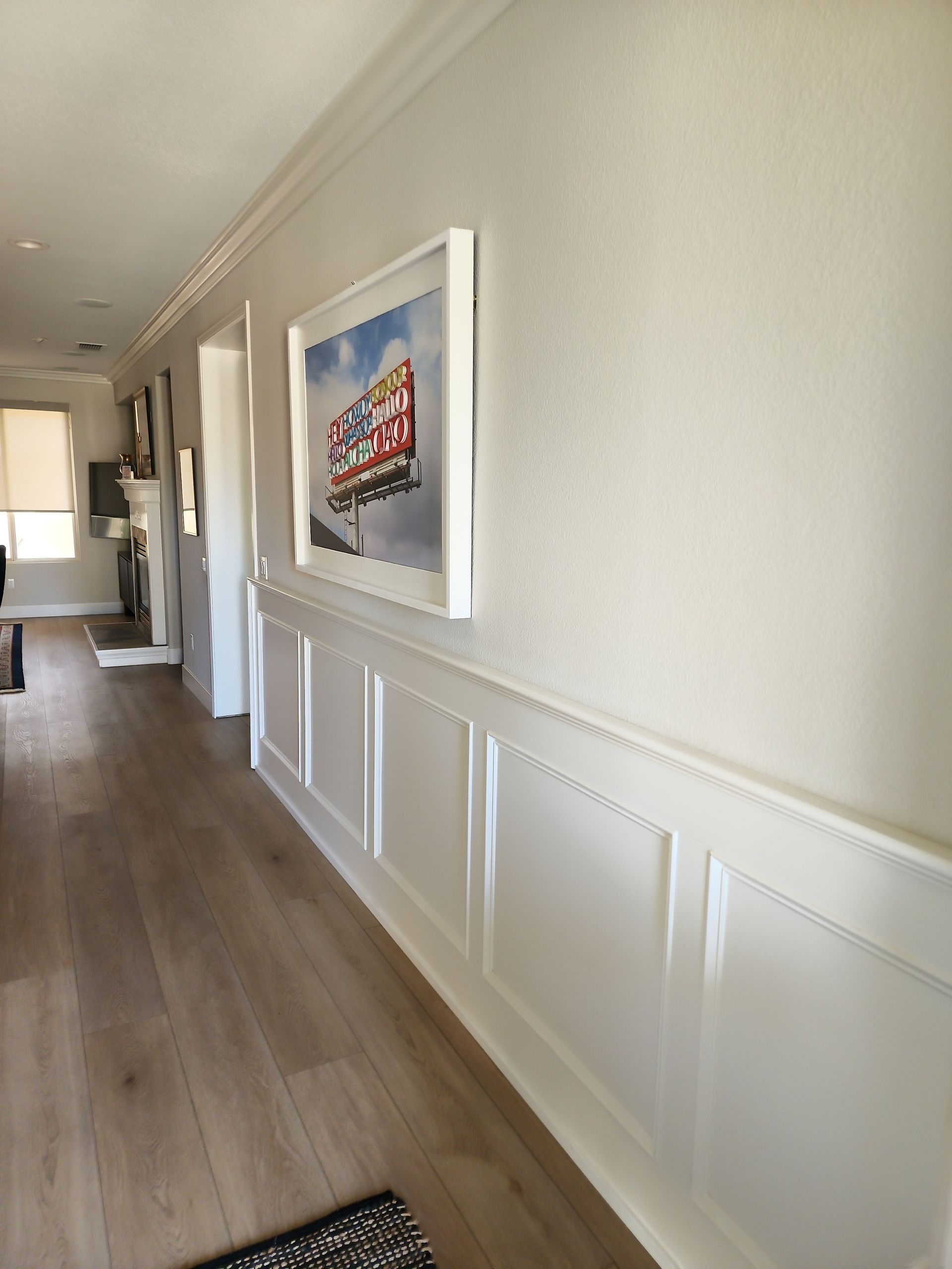 Long hallway with light wood floors and white wainscoting. A colorful framed picture hangs on the wall.