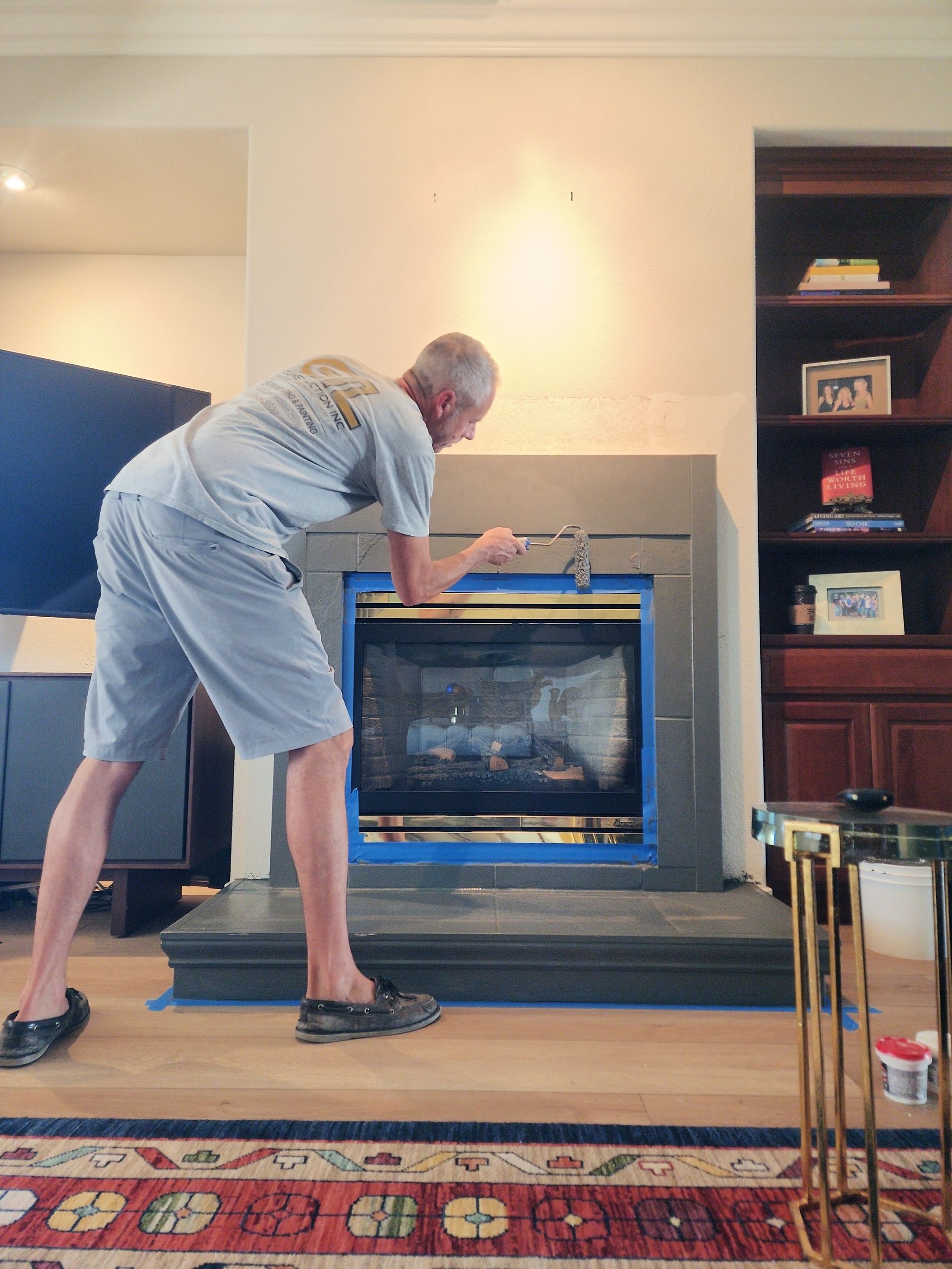 Man paints fireplace, blue tape outlines the firebox, interior setting.