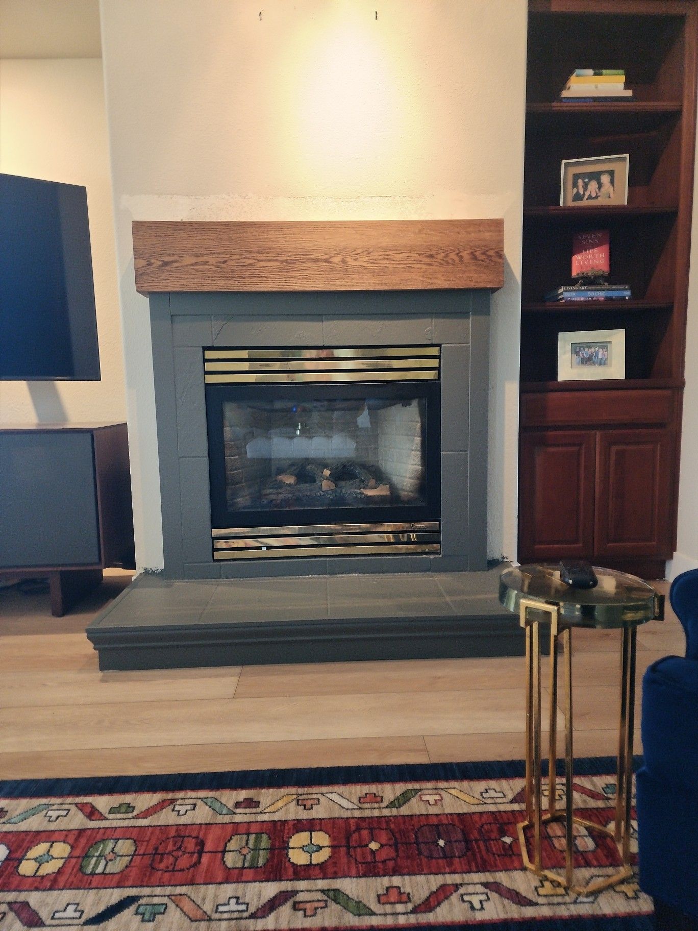 Fireplace with gray surround and wooden mantel, flanked by a TV and built-in bookshelf.