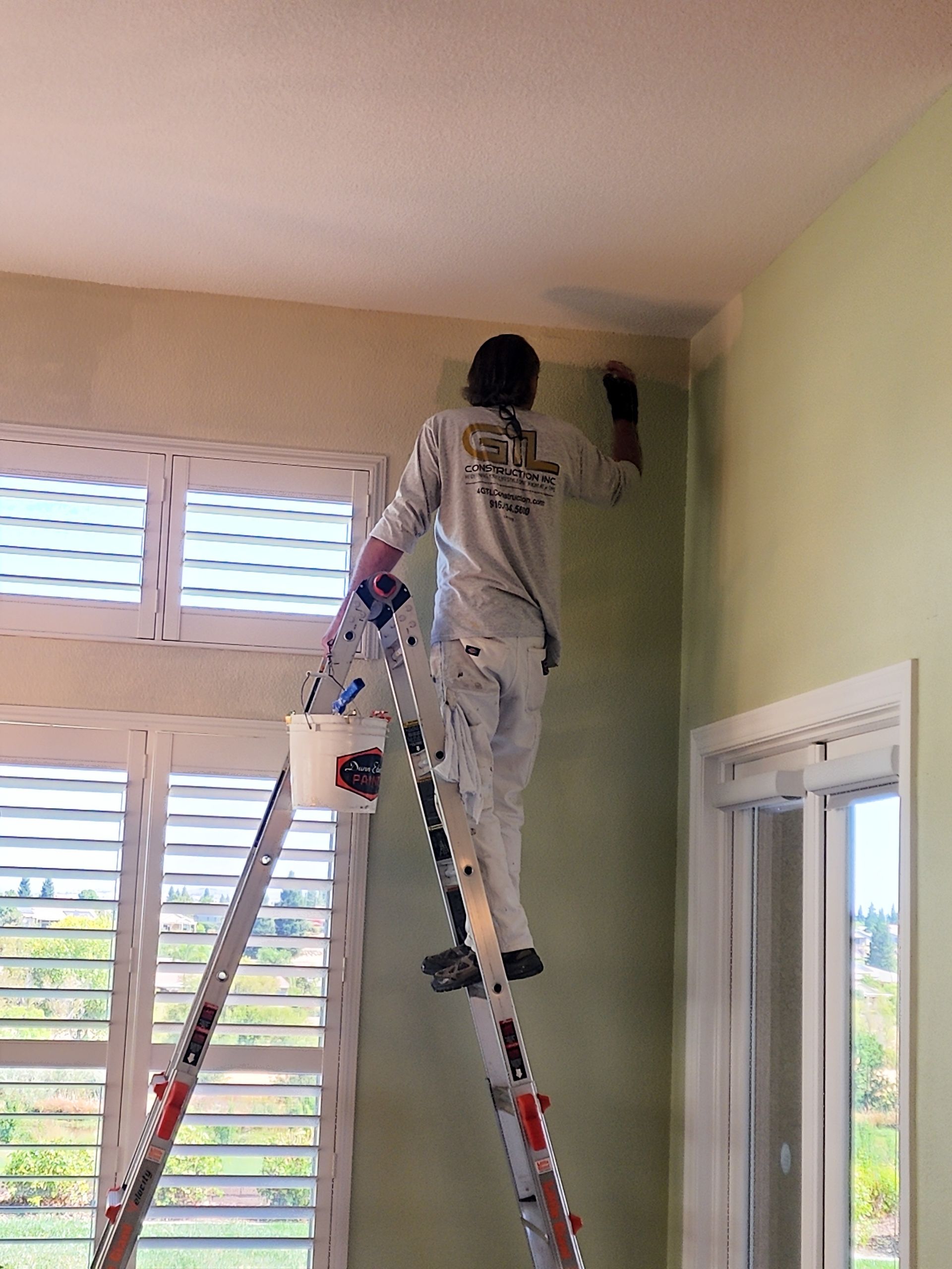 Painter on a ladder, painting trim on a green wall, with a light green ceiling and window shutters.