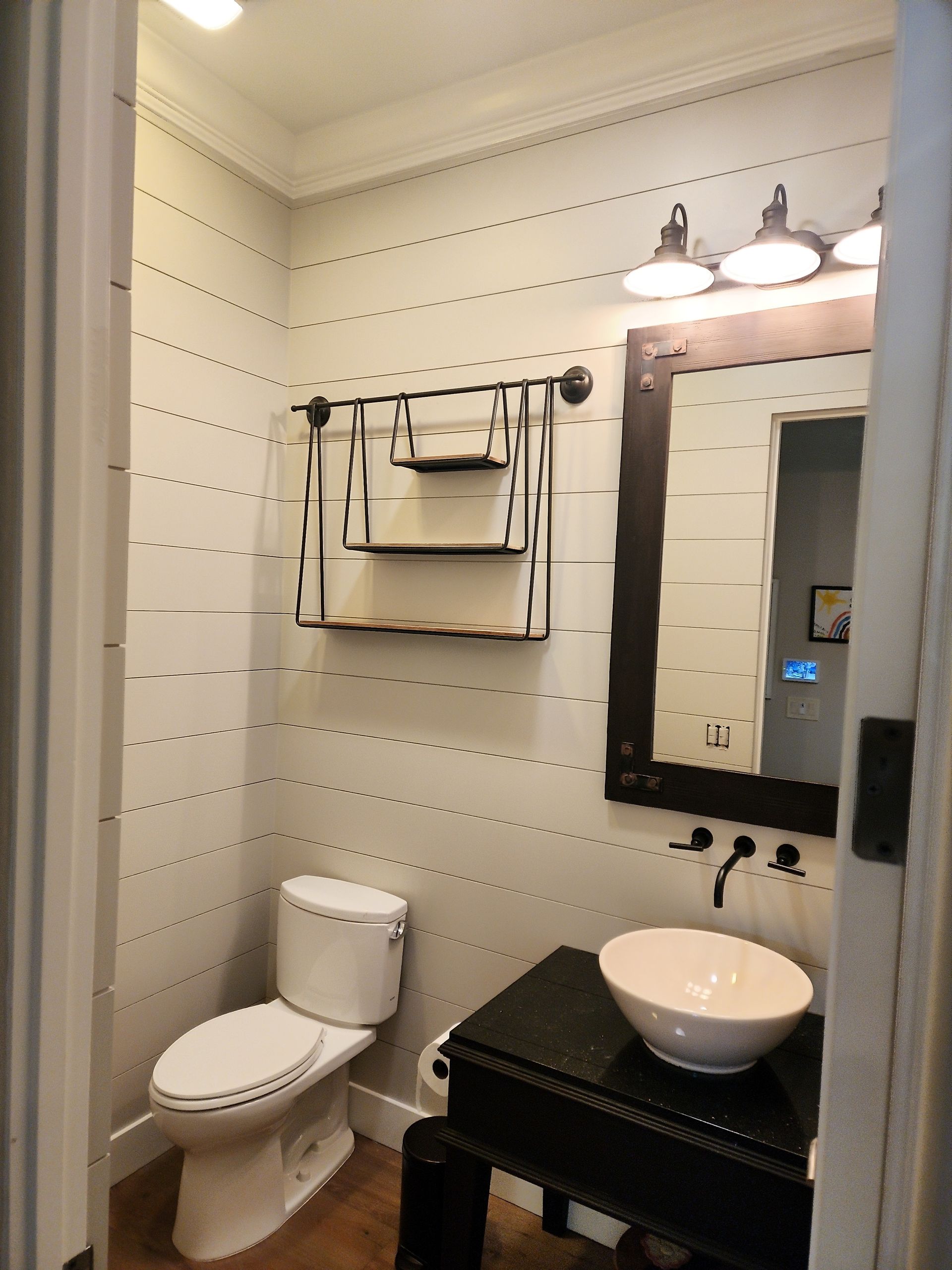 A small bathroom with a toilet, sink, mirror, and shelves. White walls with horizontal black lines.
